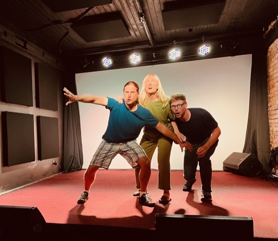 Photograph of three people on an indoor stage. One is pointing and they all look shocked.