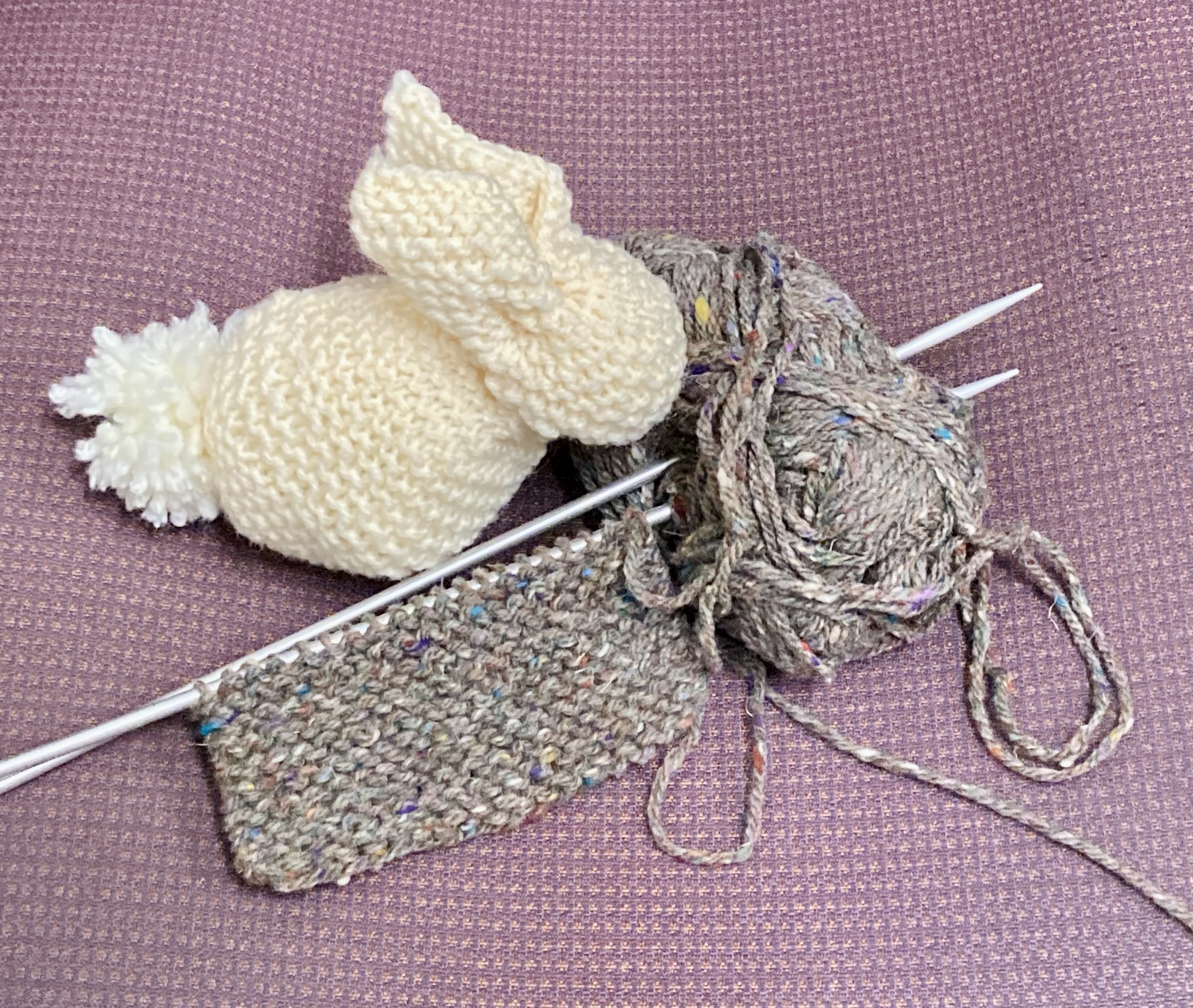 A cream colored knit bunny sits next to a knitting project on a pair of needles stabbed into gray yarn.