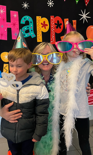 Beth poses with two young patrons in silly prop sunglasses and feathered boa.