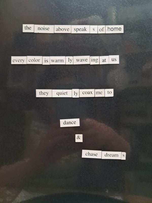 Image of magnet board poetry. The poem reads: The noise above speaks of home. Every color is warmly waving at us. They quietly coax me to dance & chase dreams.