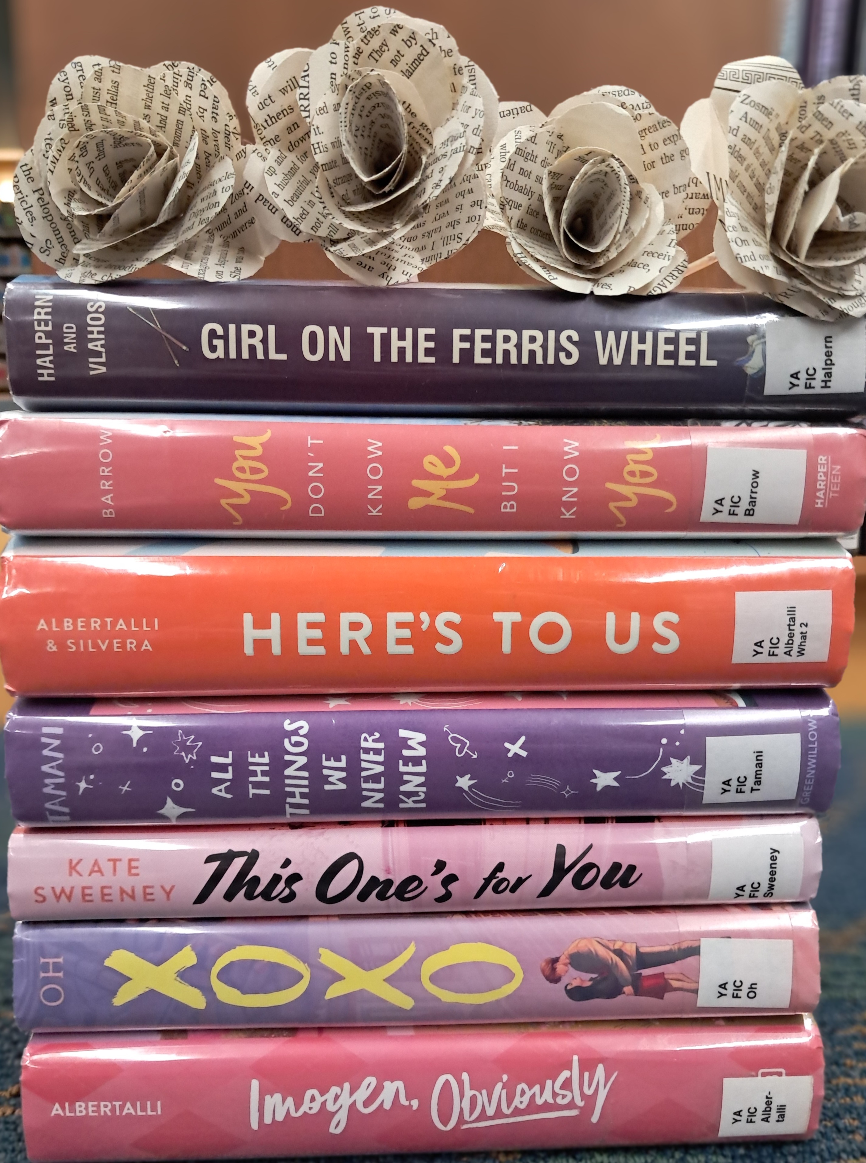 An image of book spine poetry, a form of poetry where multiple books are stacked together to form a poem. The poem is composed of 7 books, and reads: Girl on the ferris wheel, you don't know me but I know you. Here's to us, all the things we never knew. This one's for you. XOXO Imogen, obviously.