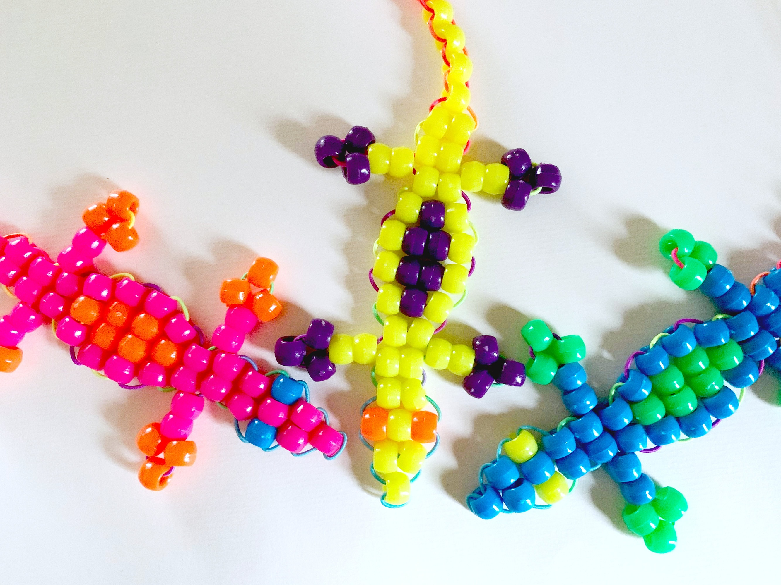 Image of 3 lizards crafted from pony beads. They are in pink and orange, yellow and purple, and blue and green respectively.