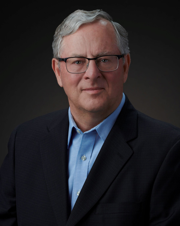 Photo of a older white man wearing glasses with black frames and short gray hair who is wearing a light blue dress shirt and a black suit coat.