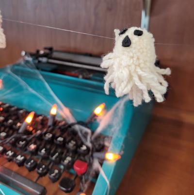 a ghost made out of yarn hangs over a web-covered teal typewriter. there are orange fairy lights on the typewriter's keys.