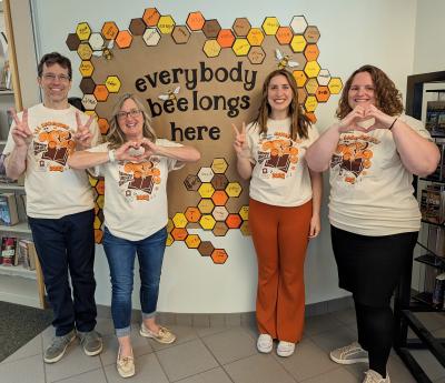 Image of four people in Summer Reading Challenge t-shirts in front of a sign that says "everyone belongs here." The four people are either showing a peace sign or making a heart with their hands.