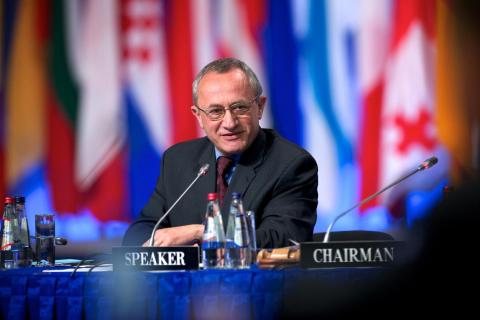 photo of an older white man sitting at a conference table with flags from various countries behind him.