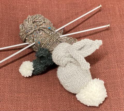 A gray and smaller green knit bunny sit next to some yarn with knitting needles stabbed through it.