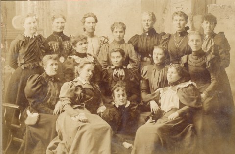 Image in sepia tones, featuring 15 women standing and sitting for a studio protrait of their group, the Ladies Club of Traverse City, from the early 1900s. Women are wearing large, sturdy dresses with full sleeves and long hems.