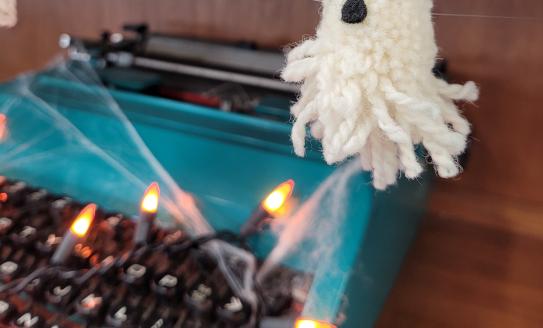 a ghost made out of yarn hangs over a web-covered teal typewriter. there are orange fairy lights on the typewriter's keys.