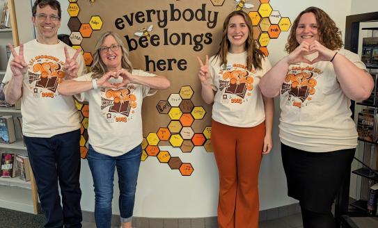 Image of four people in Summer Reading Challenge t-shirts in front of a sign that says "everyone belongs here." The four people are either showing a peace sign or making a heart with their hands.