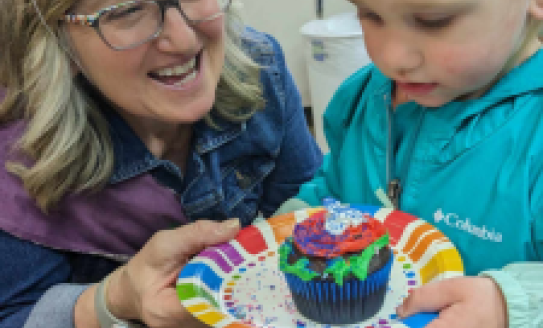 Beth and young patron in party hats smile at a brightly frosted cupcake.